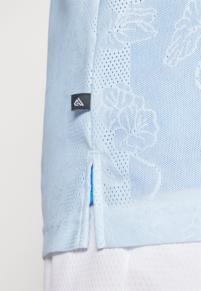 Chemise légère bleu clair à manches courtes avec un motif floral, fabriquée en tissu en maille respirant, dotée d'une fente sur le côté et d'une étiquette logo noire au bas.
