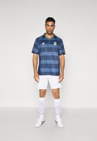 Maillot de football rayé marine avec des accents jaunes, à manches courtes et en V ; associé à un short blanc et à des chaussettes blanches montantes.