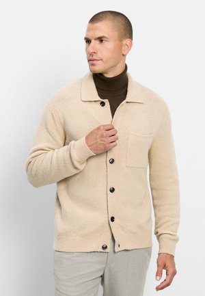 Mann trägt einen beigen, geknöpften Cardigan über einem dunkelbraunen Rollkragenpullover und schaut zur Seite vor einem einfarbigen hellen Hintergrund.