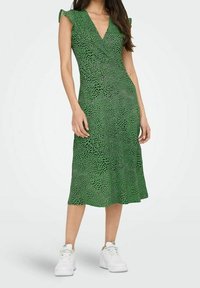 Femme portant une robe midi sans manches à col en V verte avec un motif abstrait noir, associée à des baskets blanches, se tenant devant un fond blanc.