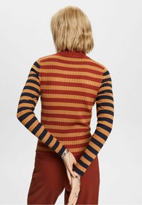 Personne blonde aux cheveux courts et ondulés, portant un pull côtelé à rayures couleur rouille, orange et marine, les mains jointes dans le dos.