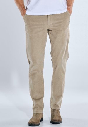 Beige Cordura-Hosen mit geradem Schnitt, ausgestattet mit Seitentaschen und einer subtilen Rippstruktur. Getragen mit braunen Wildlederstiefeln.