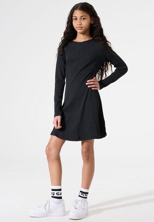 Robe noire à manches longues avec un col rond, un corsage ajusté et une jupe évasée. Associée à des baskets blanches et des chaussettes à motifs.