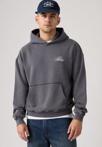 Γκρι βαμβακερό φούτερ με κουκούλα και μπροστινή τσέπη, με λευκό λογότυπο "Levi Strauss". Συνδυασμένο με μπλε τζιν παντελόνι για ένα casual look.