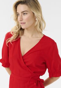 Robe enveloppante rouge avec un décolleté en V, des manches courtes bouffantes, un tissu doux et une ceinture nouée à la taille. Texture lisse, sans motifs visibles ni éléments métalliques.