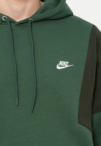 Vihreä huppari pehmeästä kankaasta, jossa on kahden sävyn suunnittelu: tummempi vihreä sivupaneeli ja valkoinen Nike-logo rinnassa.