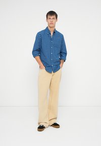 Camisa de cuadros azul y blanca con botones, pantalones beige de pierna ancha y zapatos negros con detalles en blanco. El modelo está de pie con las manos en los bolsillos.