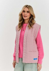 Różowa pikowana kamizelka z teksturowanym wzorem, z przodu otwarta, z dwoma bocznymi kieszeniami, założona na jasnoróżowy sweter. Widoczna niebieska metka marki.