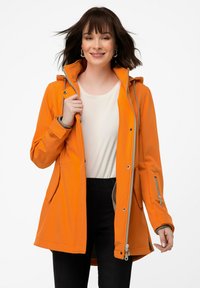 Oranssi vedenpitävä takki hupulla, jossa on vetoketjulliset taskut, hopeiset yksityiskohdat ja pehmeä sisävuori. Halkaistuna kevyen paidan päällä.