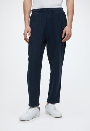 Mand iført navy blå casual bukser og hvide sneakers, stående mod en ensfarvet lys baggrund.