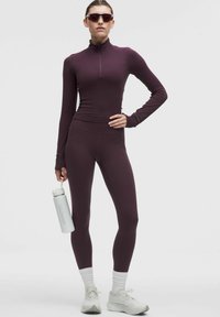 Bordeaux športni set z zip-up dolgim rokavom in visokim pasom na pajkicah. Model drži belo plastenko, nosi sončna očala in bele čevlje.