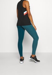 Czarny bezrękawnik z diagonalnym paskiem w kolorach czerwonym, białym i niebieskim; turkusowe legginsy; czarne sneakersy z białymi akcentami; gładki, dopasowany materiał.