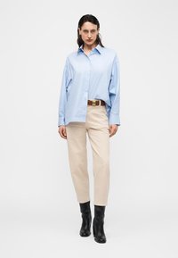 Γυναίκα που φοράει φαρδιά ανοιχτόμπλε πουκάμισο με κουμπιά, μπεζ παντελόνι με καφέ ζώνη και μαύρα μποτάκια στον αστράγαλο, στέκεται μπροστά σε λευκό φόντο.