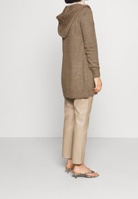 Hnědý pletený kardigan s kapucí, zdobený texturovaným vzorem a žebrovanými manžetami, kombinovaný s béžovými koženými kalhotami a sandály na řemínky.