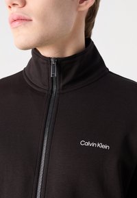 Melna zip-up jakas, kas izgatavota no mīksta auduma, ar augstu apkakli, sudraba rāvējslēdzēju un balto "Calvin Klein" logo uz krūškurvja.