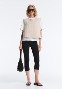 Femme portant un gilet matelassé beige oversize sur une chemise blanche, des leggings noirs courts, des tongs noires, tenant un sac noir et des lunettes de soleil noires.