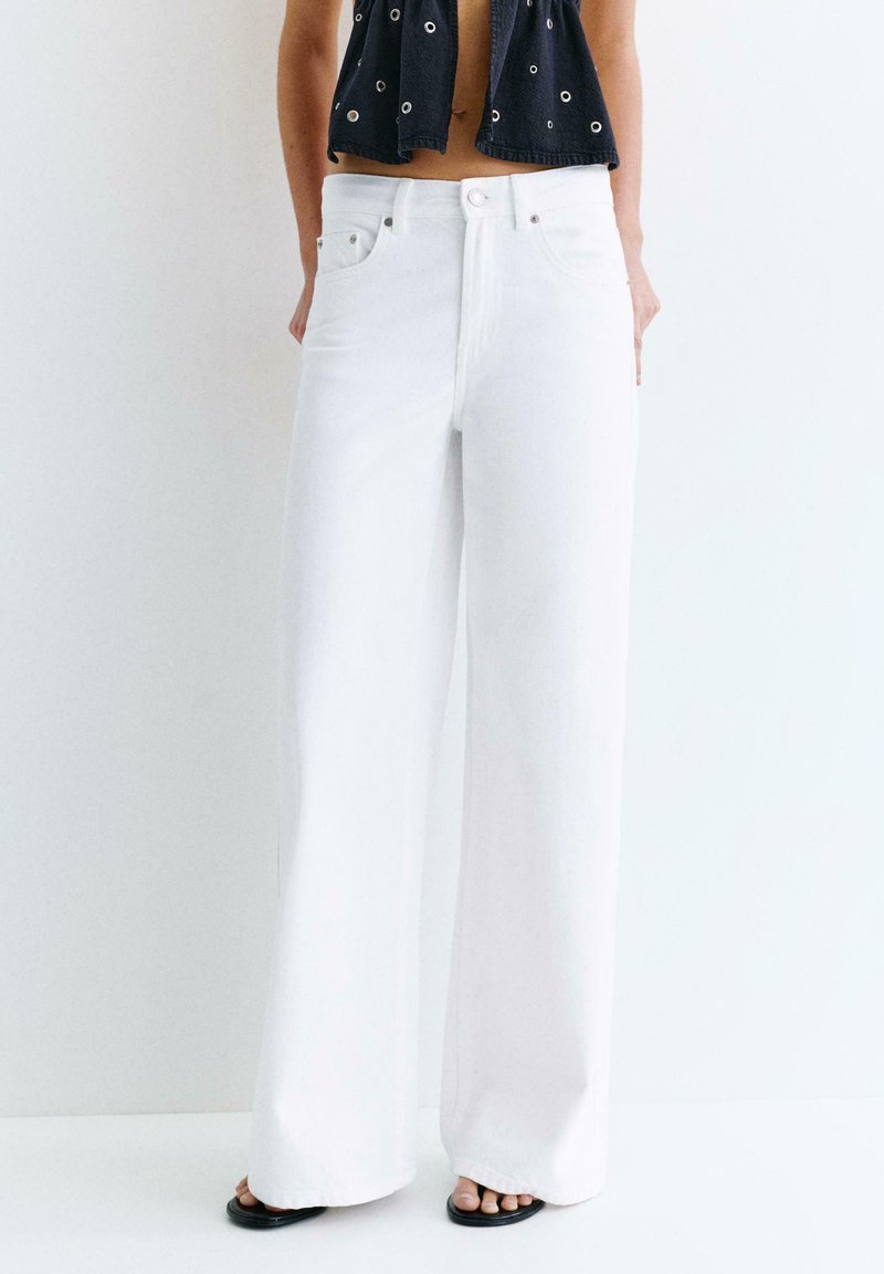 Personne portant un jean blanc taille haute à jambes larges, associé à des sandales noires à bout ouvert et un top court noir avec des détails œillets.