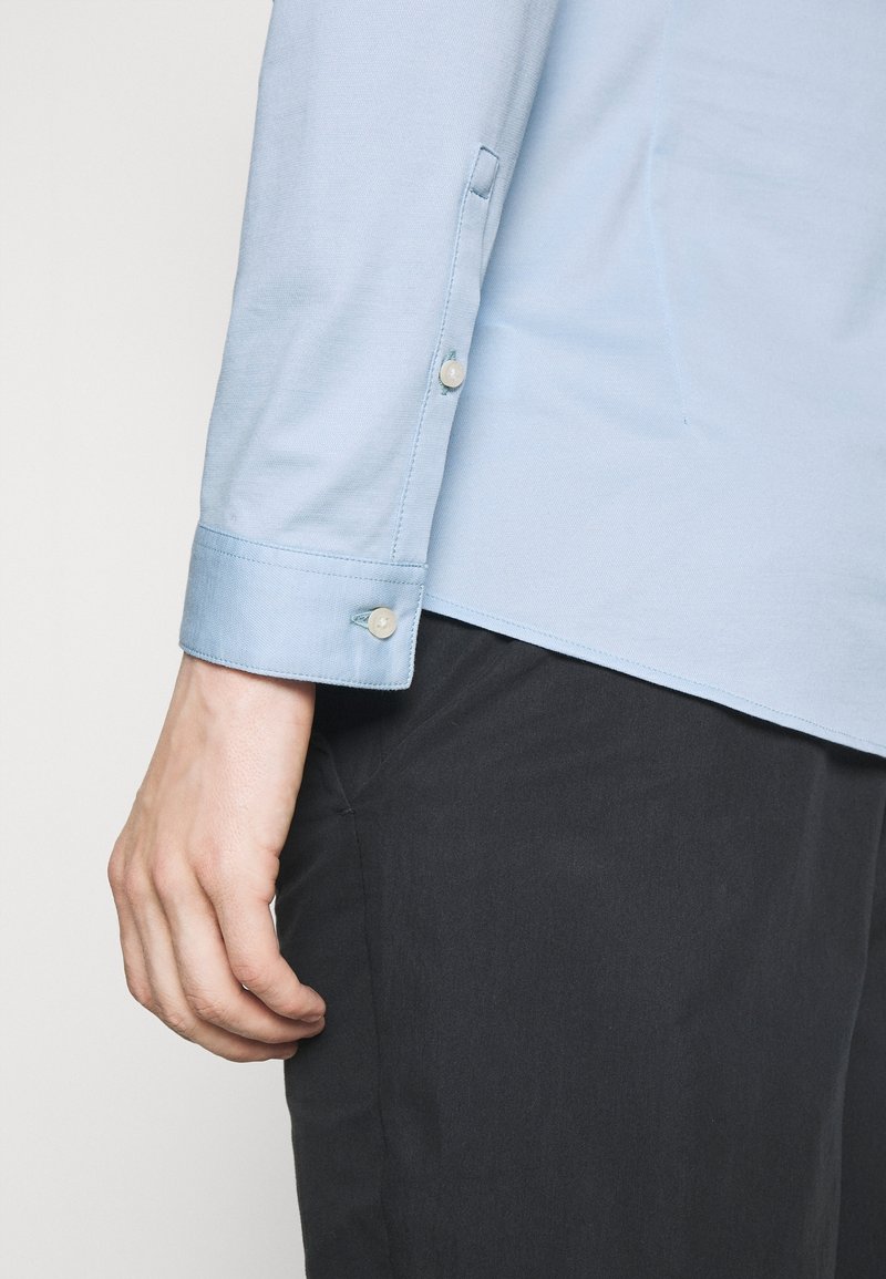 Camicia a maniche lunghe blu chiaro con polsini abbottonati, in morbido cotone e orlo dritto, abbinata a pantaloni scuri.