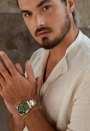 Silver and gold metal watch with a green dial on a wrist, paired with a gold chain bracelet. Light beige linen shirt with a collar.