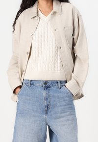 Mujer con una chaqueta beige sobre un suéter de punto en color crema y unos jeans de cintura alta en azul claro, con las manos en los bolsillos.