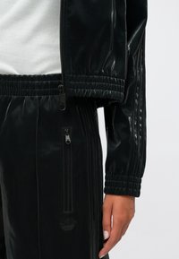 Μαύρο γυαλιστερό κοστούμι αθλητικών ρούχων από λαμπερό ύφασμα. Περιλαμβάνει ριμπ μανίκια, τσέπες με φερμουάρ και ανάγλυφο λογότυπο στα παντελόνια.