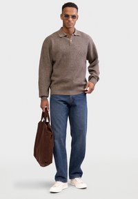 Καφέ πλεκτό πουλόβερ με υφή και γιακά πόλο, συνδυασμένο με μπλε τζιν παντελόνι. Η εμφάνιση ολοκληρώνεται με καφέ τσάντα tote και λευκά αθλητικά παπούτσια.