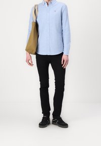 Mann trägt ein hellblaues Hemd, schwarze Hose, schwarze Turnschuhe und trägt eine braune Tragetasche mit beigen Trägern über der Schulter.