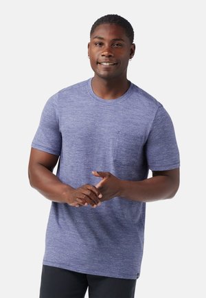 Uśmiechnięty mężczyzna o krótkich włosach, ubrany w niebieski melanżowy t-shirt z kieszonką i czarne spodnie, stojący na białym tle.