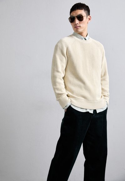 Light beige ribbed knit sweater with a crew neck over a collared light blue shirt, paired with dark, wide-leg corduroy trousers. Bronze sunglasses.