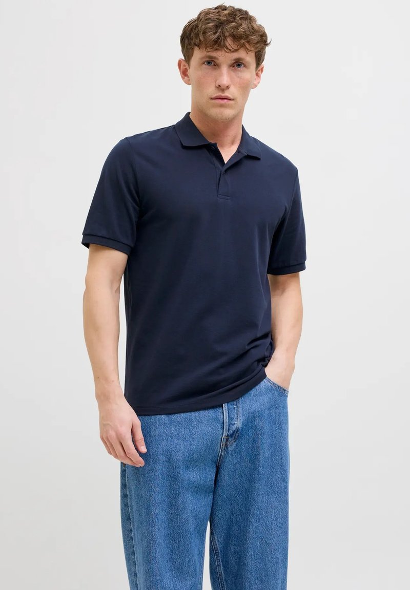 Polo shirt a manica corta blu navy con colletto, realizzato in tessuto di cotone morbido, abbinato a jeans in denim azzurro chiaro. Design semplice, vestibilità su misura.