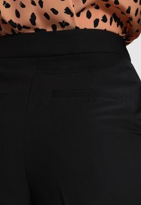 Svarta högmidjade byxor med slät yta, som har två bakfickor och en ren design, i kombination med en mönstrad persikofärgad blus.