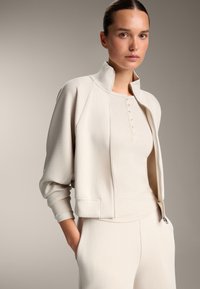 Hellbeige Sportoutfit mit einer kurvenförmigen, gepolsterten Jacke mit Rippstruktur und einem taillierten Tanktop mit Knöpfen. Glatter Stoff.