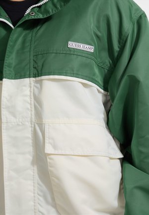 Groene en witte windbreaker met een ritssluiting, voorzien van een voorzak en het 'GUESS JEANS' logo. Glad textuur en sportief design.
