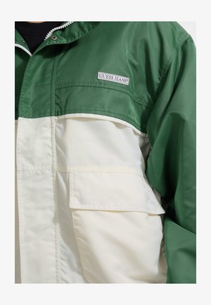 Veste coupe-vent verte et blanche avec fermeture éclair, dotée d'une poche avant et du logo 'GUESS JEANS'. Texture lisse et design sportif.