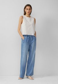 Bijela majica bez rukava s izvezenim uzorkom; uparena s svijetloplavim širokim trapericama koje imaju elastičnu struk i vezicu.