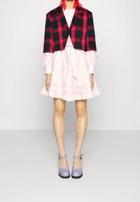 Avklippt svart och röd rutjacka över en ljusrosa klänning med volangdetaljer. Modellen bär lavendelfärgade platåskor med ankelsnören.