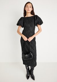 Črna obleka z rožnim vezenjem in napihnjenimi rokavi, prilega se silhueti, do kolen, v kombinaciji z majhno črno torbico in škornji do kolen s koničastim nosom.