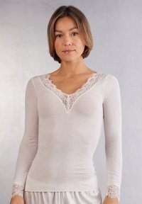Camiseta de manga larga en suave tela beige clara, con un escote en V ribeteado de encaje y acentos de encaje en los puños. Textura suave, diseño ajustado.