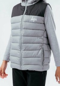 Grå vadderad väst med svarta detaljer, dragkedja framtill och sidofickor. Har en texturerad yta och en logotyp på bröstet.