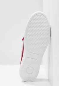 Semelle en caoutchouc blanc d'une basket rouge appuyée contre un mur blanc d'angle, montrant une empreinte texturée avec des motifs circulaires.