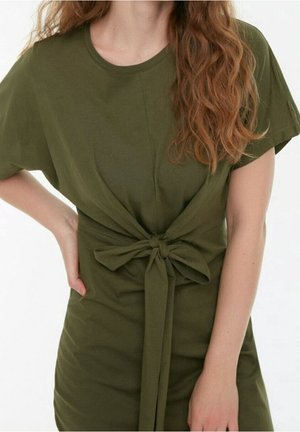 Frau trägt olivegrünes, kurzärmeliges Kleid mit Stoffgürtel, der in einer Schleife an der Taille gebunden ist, Hand ruht auf der Hüfte.