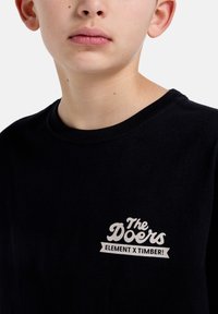Černé bavlněné tričko s bílým potiskem na prsou, označené "The Doers ELEMENT x TIMBER!" s neformálním kulatým výstřihem.