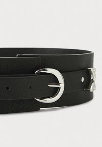 Cinturón de cuero negro con un acabado texturizado, que presenta una hebilla circular plateada y ajustes de múltiples agujeros para el tamaño.