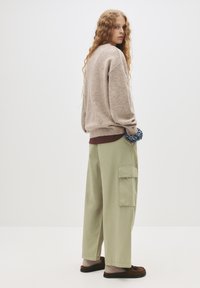 Jasnobrązowy, oversize sweter, zielone, szerokie spodnie cargo z bocznymi kieszeniami oraz brązowe buty wsuwane. Gładka tekstura, luźna sylwetka.