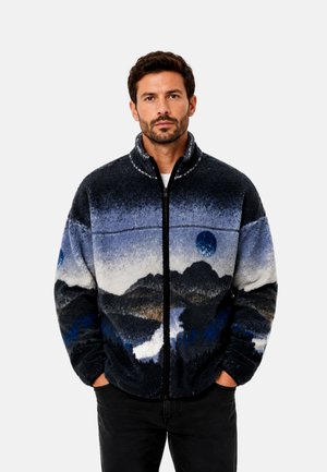 Homme portant une veste en polaire zippée avec un motif paysage de montagnes et lune bleu et noir, mains dans les poches, fond neutre.