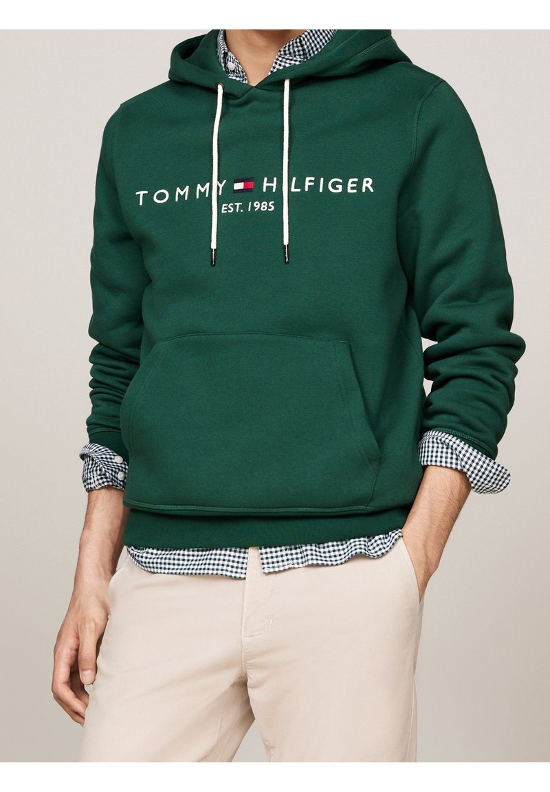 Zelená mikina z bavlny s přední kapsou, bílými šňůrkami a nápisem loga. Přes ní je vrstvená modro-bílá kostkovaná košile.