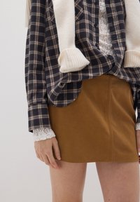Camicia a quadri navy e tan, indossata sopra un top bianco in pizzo, abbinata a una gonna in suede marrone. Presenta maniche a coste color crema e dettagli sulle cuciture laterali.