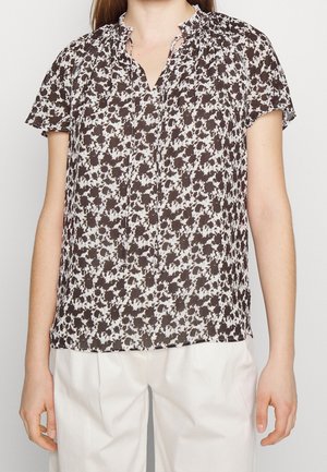 Vrouw draagt een zwart-wit bloemenblouse met korte mouwen en een geplooide halslijn, gecombineerd met een witte broek, staand tegen een effen achtergrond.