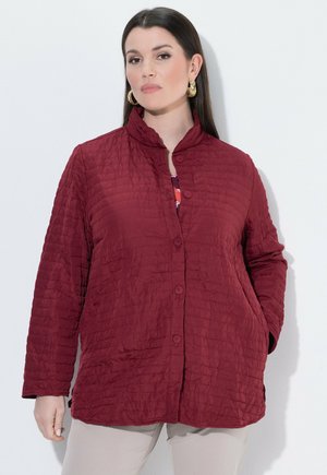 Femme portant une veste matelassée bordeaux avec des boutons, debout devant un fond clair uni, mains dans les poches.