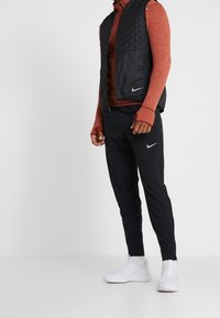 Person in schwarzen Nike Jogginghosen, weißen Nike Sneakern, einer schwarzen gesteppte Weste und einem rostfarbenen Langarmshirt, stehend auf einem weißen Boden.